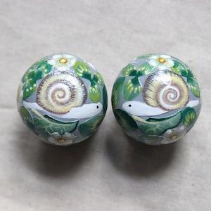 May include: Two round, hand-painted decorative objects. Each features a snail with a cream-colored shell, surrounded by green leaves and white flowers, set against a gray background. The snail's body is white.