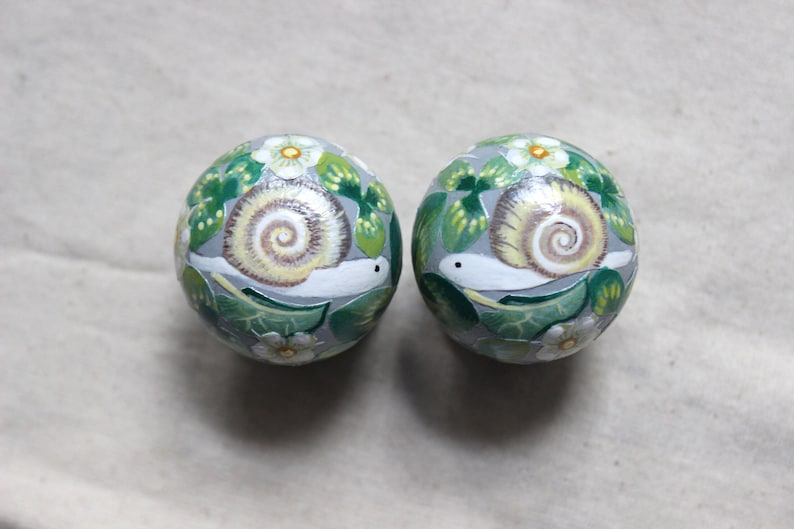 May include: Two round, hand-painted decorative objects. Each features a snail with a cream-colored shell, surrounded by green leaves and white flowers, set against a gray background. The snail's body is white.
