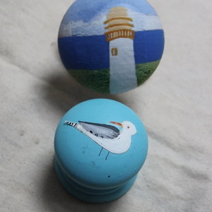 May include: Two painted wooden knobs. One is light blue with a white seagull design. The other is painted with a lighthouse scene, featuring a white tower, tan top, blue sea, and sky.