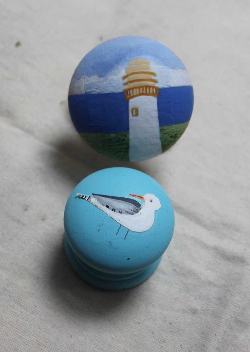 May include: Two painted wooden knobs. One is light blue with a white seagull design. The other is painted with a lighthouse scene, featuring a white tower, tan top, blue sea, and sky.