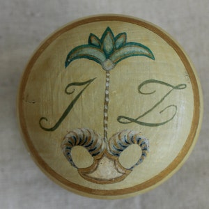 May include: A round wooden ornament with a gold border and a painted design. The design features a stylised green palm tree with a white trunk and a gold and blue swirl at the base. The letters "IZ" are painted in green on either side of the tree.