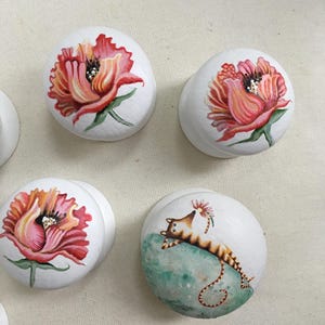 May include: White cabinet knobs with hand-painted floral and cat designs. The knobs feature vibrant red, pink, and orange flowers, and a whimsical cat resting on a green surface. These decorative knobs add a unique touch to furniture.