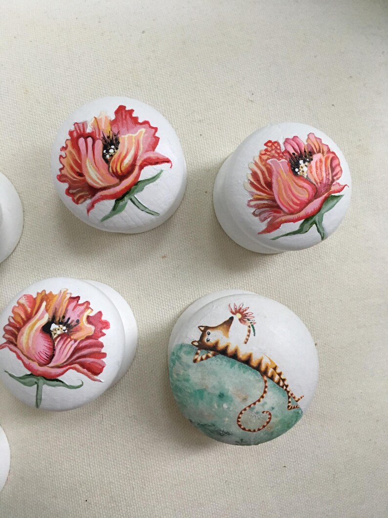 Puede incluir: Pomos de armario blancos con dise&ntilde;os florales y de gatos pintados a mano. Los pomos presentan vibrantes flores rojas, rosas y naranjas, y un gato caprichoso descansando sobre una superficie verde. Estos pomos decorativos a&ntilde;aden un toque &uacute;nico a los muebles.