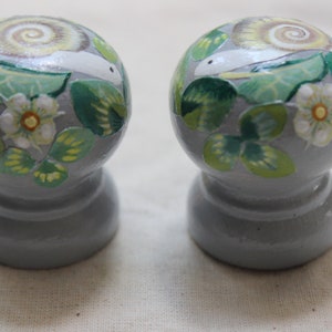 May include: Two gray, round decorative knobs with a painted design of a snail, green leaves, and white flowers. The snail's shell has a spiral pattern. The knobs are likely for furniture or decor.