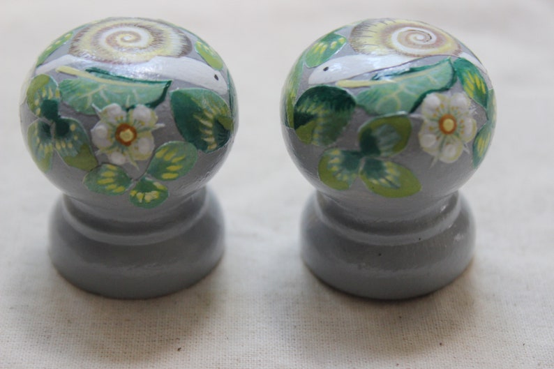 May include: Two gray, round decorative knobs with a painted design of a snail, green leaves, and white flowers. The snail's shell has a spiral pattern. The knobs are likely for furniture or decor.