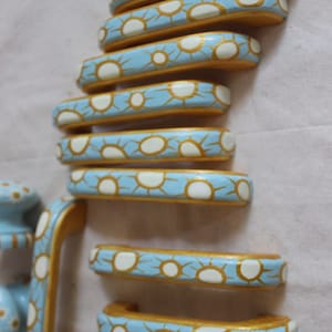 Pode incluir: Um conjunto de 10 botões de armário pintados de azul e dourado com um design de sol branco. Os botões estão dispostos em uma pilha, com o botão maior na parte inferior e o botão menor na parte superior.