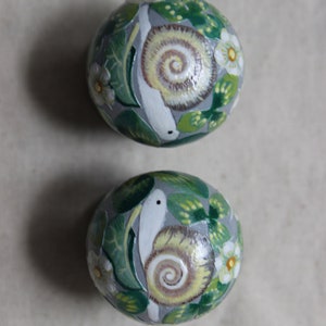 May include: Two hand-painted decorative spheres with a snail and floral design. The spheres are painted with a gray background, green leaves, white flowers, and a snail with a yellow and brown shell.