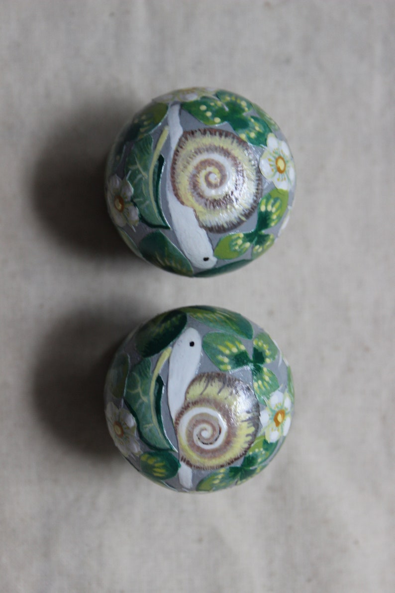 May include: Two hand-painted decorative spheres with a snail and floral design. The spheres are painted with a gray background, green leaves, white flowers, and a snail with a yellow and brown shell.