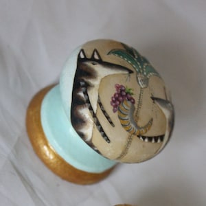 Könnte beinhalten: Ein handbemalter Holz-Knopf mit einem hellblauen Sockel und einem goldenen Rand. Die Oberseite des Knopfes zeigt eine skurrile Illustration eines gestreiften Tieres mit einem Schwanz, einer Traube und einer Pflanze mit Blättern.