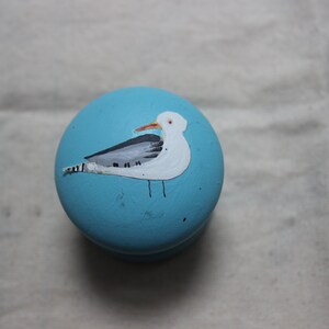 May include: A round, light blue decorative knob with a hand-painted seagull design. The seagull is white with gray wings and a red beak. The knob is likely made of wood and is suitable for furniture or decor.