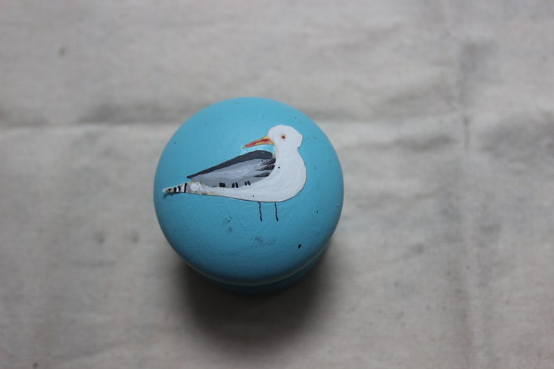 May include: A round, light blue decorative knob with a hand-painted seagull design. The seagull is white with gray wings and a red beak. The knob is likely made of wood and is suitable for furniture or decor.