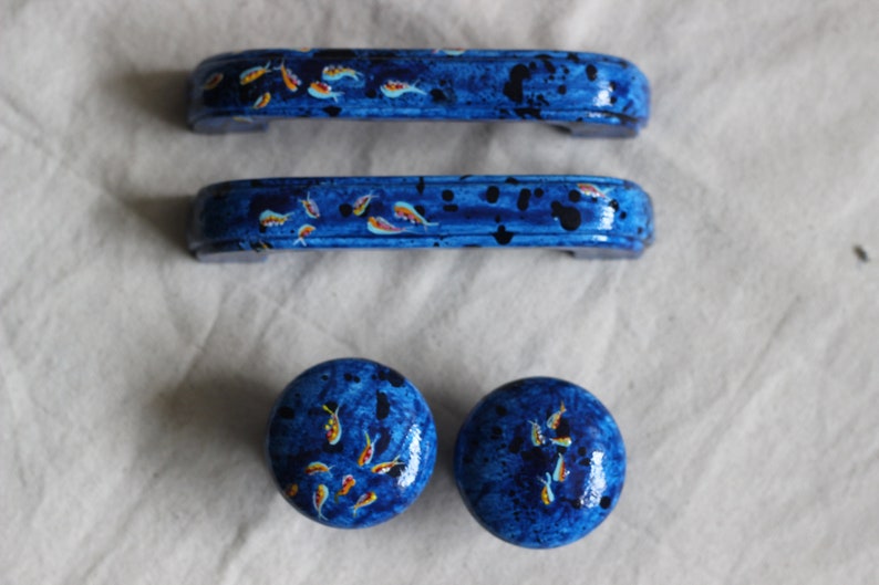 May include: Three blue cabinet knobs with a painted design of orange and yellow fish on a blue background. The knobs are round and have a slightly raised edge.
