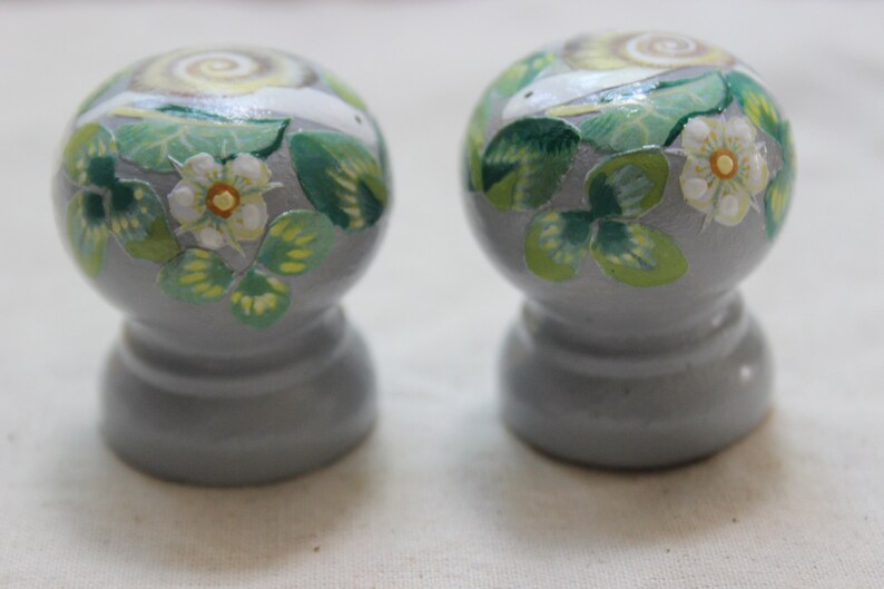 May include: Two gray, round salt and pepper shakers with a painted design. The design features a snail, white flowers, and green leaves. The shakers have a bulbous top and a pedestal base.