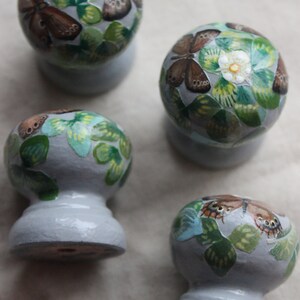 Puede incluir: Cuatro pomos de cer&aacute;mica con una base gris claro y un dise&ntilde;o de mariposa y flor blanco, verde y marr&oacute;n.