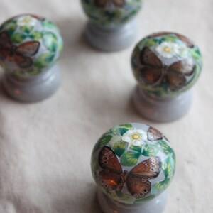 Puede incluir: Cuatro adornos redondos decorativos con fondo blanco y hojas verdes. Cada adorno presenta una mariposa marr&oacute;n con detalles blancos. Los adornos est&aacute;n sobre una superficie gris claro.