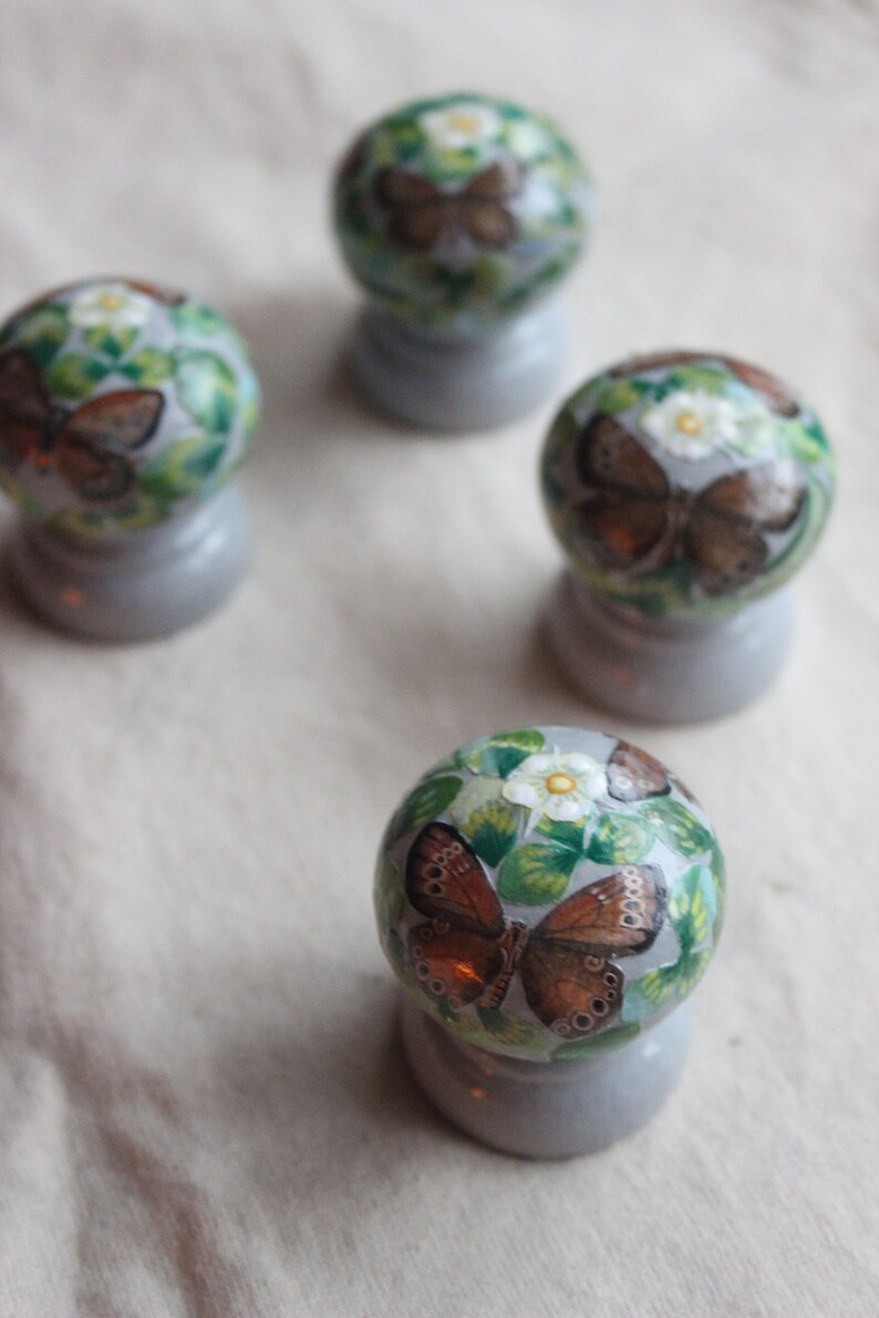 Puede incluir: Cuatro adornos redondos decorativos con fondo blanco y hojas verdes. Cada adorno presenta una mariposa marr&oacute;n con detalles blancos. Los adornos est&aacute;n sobre una superficie gris claro.