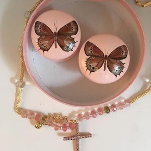 Peut inclure: Une bo&icirc;te &agrave; bijoux rose avec deux couvercles ronds, chacun orn&eacute; d'un motif de papillon peint. Un collier dor&eacute; avec une croix et des perles en cristal rose est au premier plan. La bo&icirc;te et les couvercles sont rose clair.