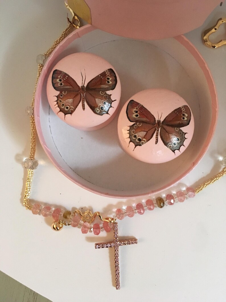 Peut inclure: Une bo&icirc;te &agrave; bijoux rose avec deux couvercles ronds, chacun orn&eacute; d'un motif de papillon peint. Un collier dor&eacute; avec une croix et des perles en cristal rose est au premier plan. La bo&icirc;te et les couvercles sont rose clair.