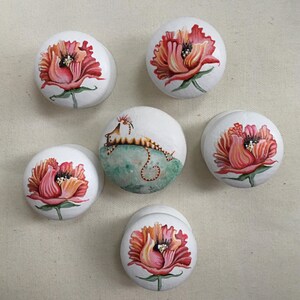 Puede incluir: Seis piedras blancas lisas pintadas a mano. Cinco piedras presentan una vibrante flor roja y rosa con tallos y hojas verdes. Una piedra tiene una ilustraci&oacute;n caprichosa de una criatura sobre un fondo verde y azul.