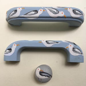 Peut inclure: Deux poignées d'armoire bleues et un bouton, tous décorés de mouettes peintes. Les poignées sont incurvées et ont un fond bleu clair. Les mouettes sont blanches avec des ailes grises et des becs orange. Le bouton est gris avec une seule mouette.