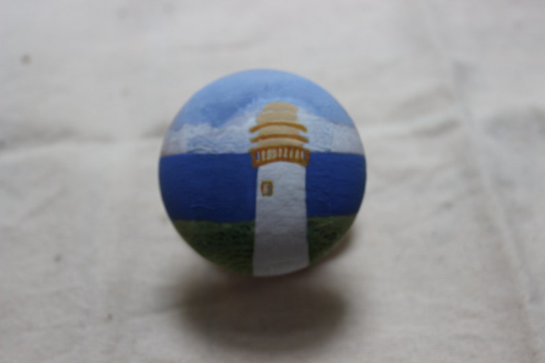 May include: A hand-painted, round decorative object featuring a lighthouse scene. The top portion depicts a blue sky with white clouds, transitioning to a blue sea. The lighthouse is white with a tan top and sits on a green base.
