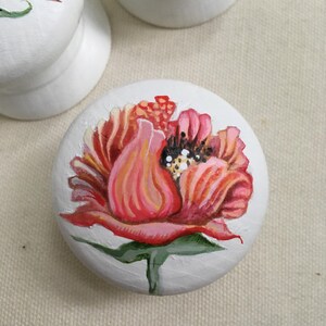 Puede incluir: Pomos de armario de madera blanca con dise&ntilde;os florales pintados a mano. Los pomos presentan una vibrante flor roja y rosa con hojas verdes. El centro de la flor tiene detalles en blanco y negro. Los pomos son redondos y tienen una base plana.