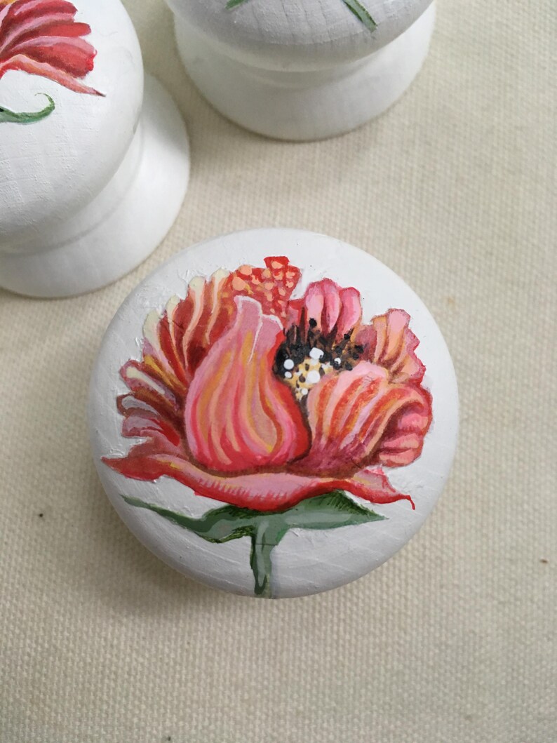 Puede incluir: Pomos de armario de madera blanca con dise&ntilde;os florales pintados a mano. Los pomos presentan una vibrante flor roja y rosa con hojas verdes. El centro de la flor tiene detalles en blanco y negro. Los pomos son redondos y tienen una base plana.