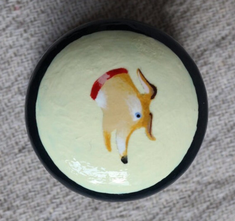 May include: A circular, hand-painted decorative item with a black border. The centre displays a light yellow backdrop with a cartoon deer illustration in tan, brown, red, and blue. The deer has a red stripe on its back.