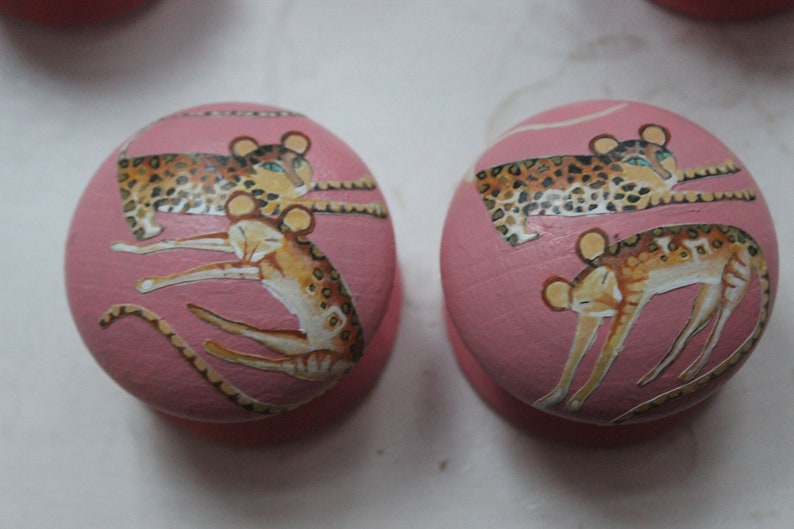 Peut inclure: Deux boutons ronds roses avec un motif de l&eacute;opards et un petit animal tachet&eacute;. Les boutons sont peints avec un fond rose et les animaux sont peints en brun, noir et blanc.