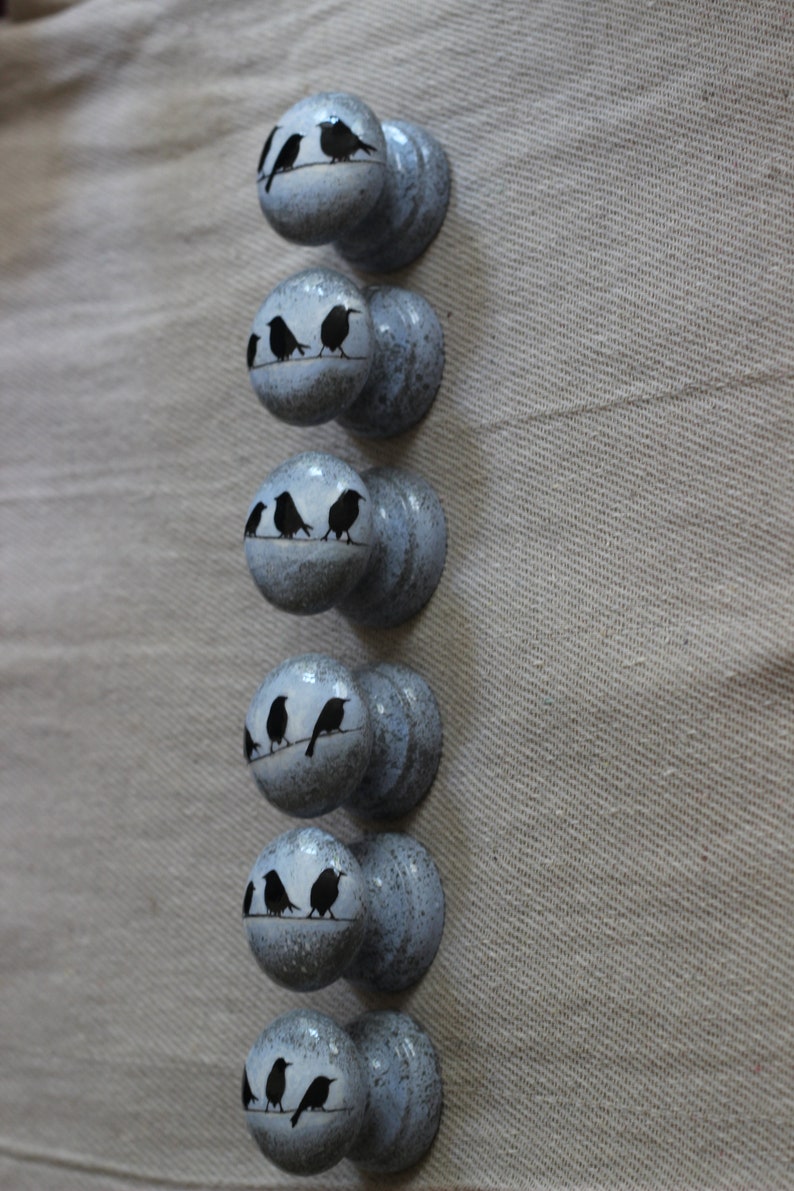 K&ouml;nnte beinhalten: Sechs blaue und graue bemalte Holzkn&ouml;pfe mit schwarzen Silhouetten von V&ouml;geln, die auf einem Ast sitzen.
