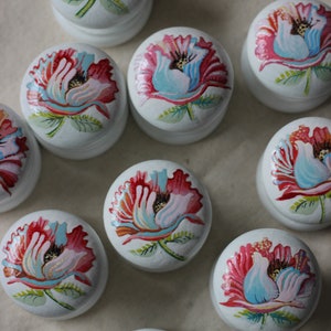 Peut inclure: Plusieurs boîtes décoratives rondes blanches, ornées de motifs floraux peints à la main. Chaque boîte présente une fleur rouge, rose et bleue avec des feuilles vertes. Les boîtes sont disposées sur une surface de couleur neutre.