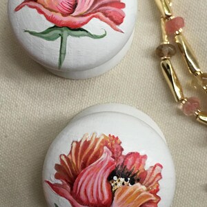 Puede incluir: Dos cajas decorativas redondas blancas con dise&ntilde;os florales pintados a mano. Las flores son en tonos de rojo, rosa y naranja, con tallos y hojas verdes. Una cadena dorada con cuentas rosas y marrones est&aacute; en el fondo.