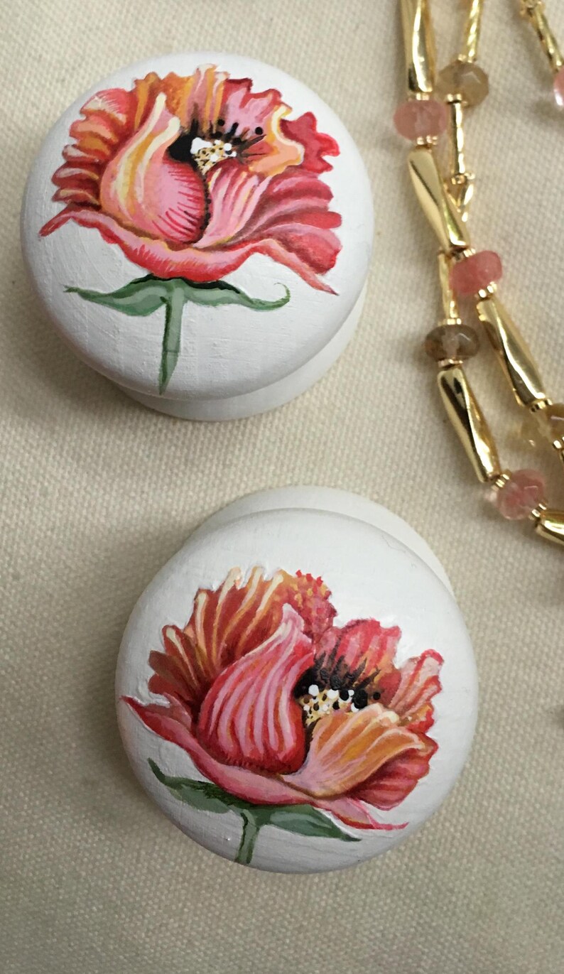 Puede incluir: Dos cajas decorativas redondas blancas con dise&ntilde;os florales pintados a mano. Las flores son en tonos de rojo, rosa y naranja, con tallos y hojas verdes. Una cadena dorada con cuentas rosas y marrones est&aacute; en el fondo.