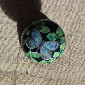 Op de afbeelding: Een handbeschilderde, ronde, zwarte steen met een gedetailleerde vlinder met blauwe en paarse vleugels. De vlinder is omgeven door groene bladeren. Een decoratief item.