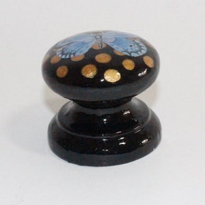 Pode incluir: Bot&atilde;o de madeira preto com uma borboleta azul e bolinhas douradas pintadas no topo.