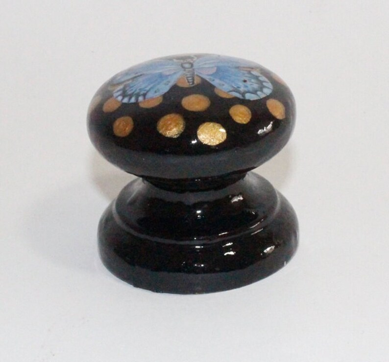 Pode incluir: Bot&atilde;o de madeira preto com uma borboleta azul e bolinhas douradas pintadas no topo.