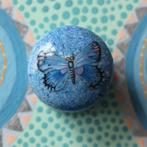 Puede incluir: Un pomo de cerámica azul con un diseño de mariposa pintado. El pomo tiene un acabado azul moteado y una mariposa blanca con contornos negros.