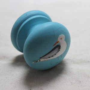 May include: A light blue, hand-painted wooden knob with a white seagull design. The knob has a rounded shape with a wider base and a narrower top. The seagull is depicted with a white body, gray wings, and an orange beak.