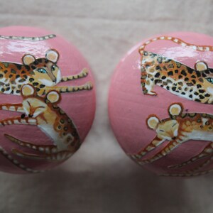 Peut inclure: Deux ornements ronds roses avec des images peintes de l&eacute;opards et de petits animaux. Les l&eacute;opards sont bruns avec des taches noires et ont des yeux bleus. Les petits animaux sont bruns avec des rayures noires et ont le ventre blanc.