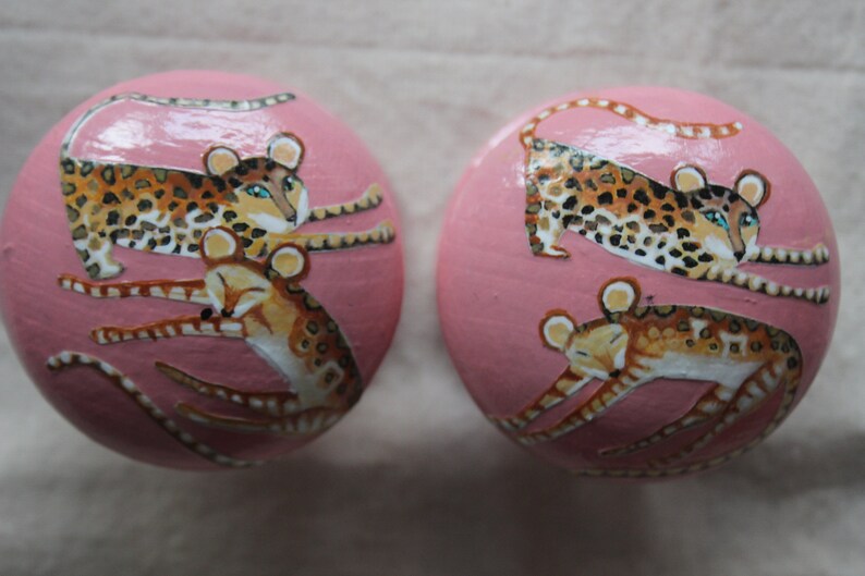 Peut inclure: Deux ornements ronds roses avec des images peintes de l&eacute;opards et de petits animaux. Les l&eacute;opards sont bruns avec des taches noires et ont des yeux bleus. Les petits animaux sont bruns avec des rayures noires et ont le ventre blanc.
