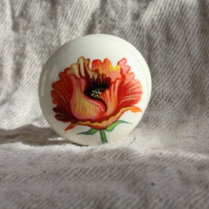 Pode incluir: Puxador decorativo redondo branco com um design detalhado de uma flor de papoula vermelha e laranja. O design floral está centrado no puxador. O puxador é provavelmente feito de cerâmica ou porcelana.