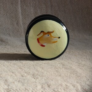 May include: A circular decorative item, black and yellow, featuring a painted dog portrait. The dog has orange fur, blue eyes, and a red collar. It could be a cabinet knob or drawer pull.