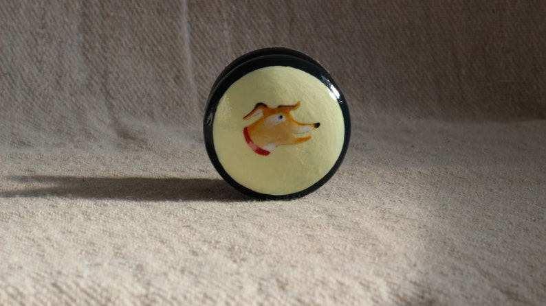 May include: A circular decorative item, black and yellow, featuring a painted dog portrait. The dog has orange fur, blue eyes, and a red collar. It could be a cabinet knob or drawer pull.