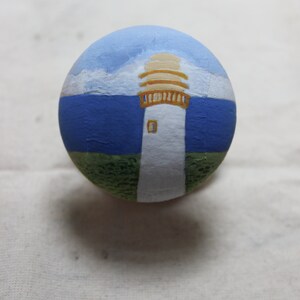 May include: A hand-painted, round stone featuring a lighthouse scene. The lighthouse is white with a tan top, set against a blue sky and sea. Green grass is at the base of the lighthouse.