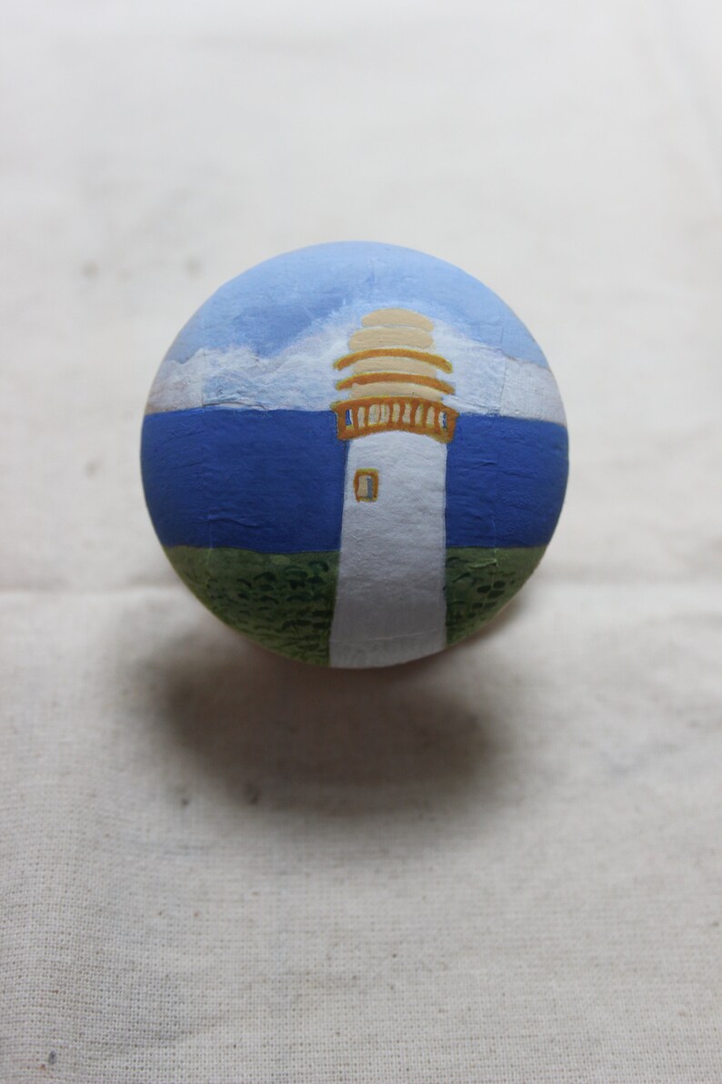 May include: A hand-painted, round stone featuring a lighthouse scene. The lighthouse is white with a tan top, set against a blue sky and sea. Green grass is at the base of the lighthouse.