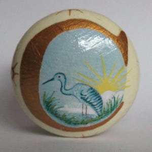 Könnte beinhalten: Runder, cremefarbener dekorativer Knopf mit einem bemalten Design. Das Design zeigt einen blauen Reiher, der in grünem Gras steht, mit einem gelben Sonnenaufgang in einem hellblauen Himmel. Ein goldener Rand umgibt das zentrale Bild.