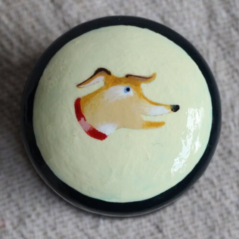 Puede incluir: Un objeto decorativo redondo y pintado con la cabeza de un perro de dibujos animados. El perro tiene pelaje marr&oacute;n claro, ojos azules y un collar rojo. El fondo es amarillo claro, con un borde negro.