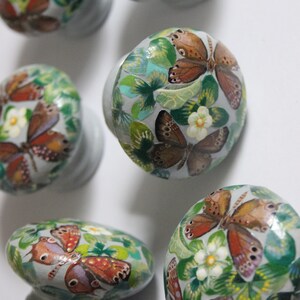 Puede incluir: Cinco pomos decorativos para armarios con base gris y dise&ntilde;o pintado de mariposas y hojas verdes. Las mariposas son marrones, naranjas y blancas.