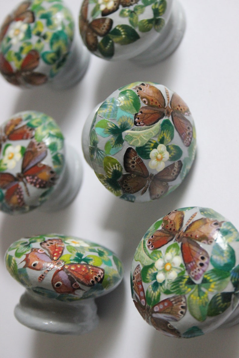 Puede incluir: Cinco pomos decorativos para armarios con base gris y dise&ntilde;o pintado de mariposas y hojas verdes. Las mariposas son marrones, naranjas y blancas.
