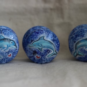Puede incluir: Tres pomos de cerámica azul y blanco con un diseño de delfín. Cada pomo presenta un delfín azul saltando fuera del agua con un corazón rojo en el fondo. Los pomos son perfectos para agregar un toque náutico a cualquier habitación.